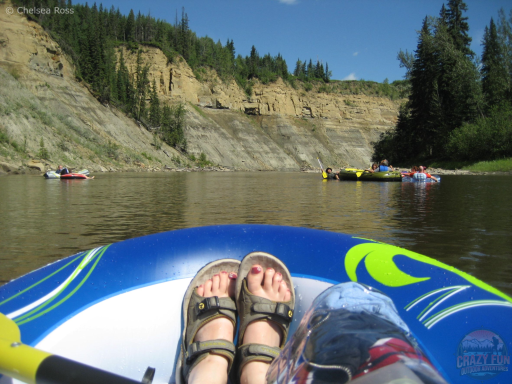 A picture of my feet in sandals in the raft with my clear bag holding my contents to keep them waterproof. Groups of rafters are in front on the calm waters. 