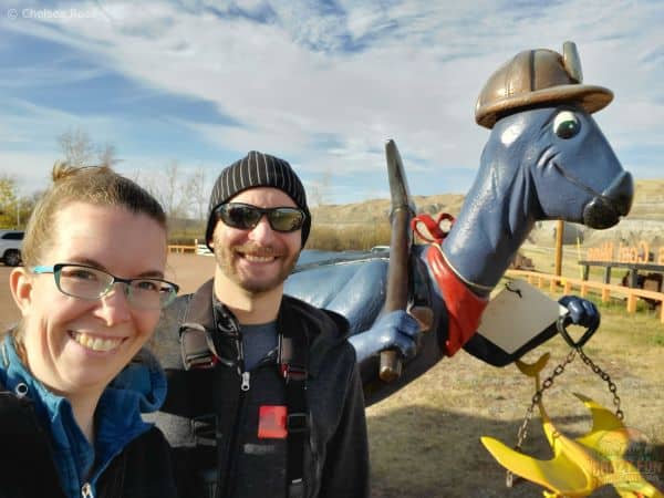 A selfie of two people with a blue dinosaur in the background enjoying Drumheller hikes.