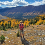 The best larch hikes showcasing my mom at Little Arethusa with beautiful larches and a mountain behind her.