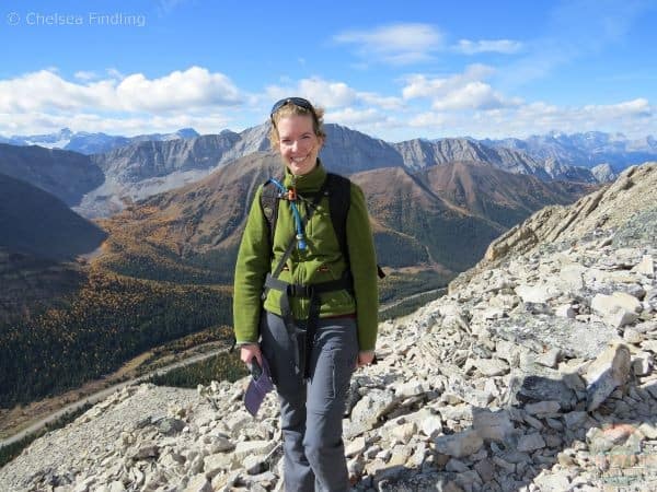 Lady wearing a green fleece and grey pants is standing on the summit with mountains and larches behind her.