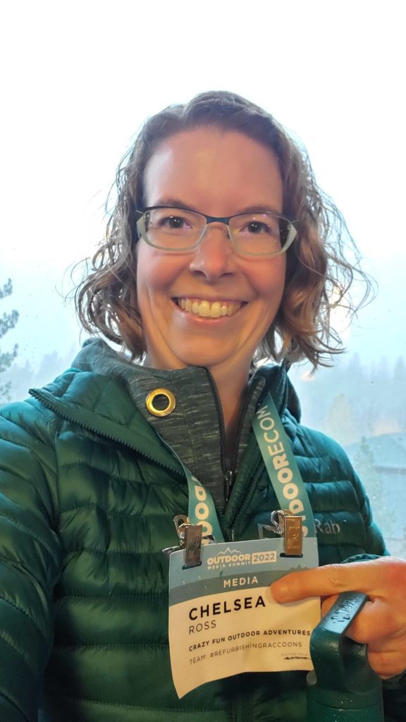 Taking a selfie at the Outdoor Media Summit while showing off my lanyard with my name on it. I'm wearing a green coat and holding a water bottle.