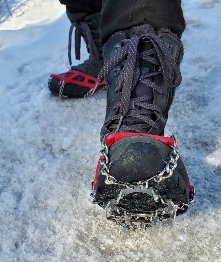 Showing a close-up of the Kahtoola Microspikes sitting underneath the black boot to the right. To the left the red stretchy part can be seen sitting on the boot.