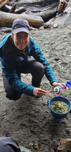 Sitting down on my feet making supper. I'm pouring dehydrated chicken and veggies into my pot. I'm wearing a blue coat and there is sand around me.