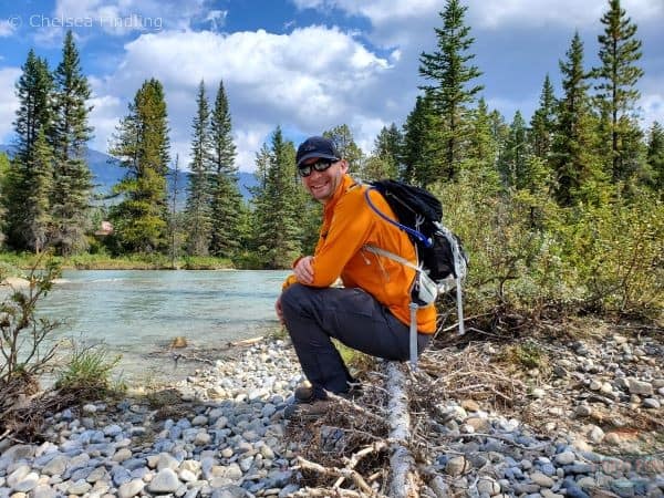 Man in a yellow fleece with an osprey backpack on is back. A river flows to his left with trees and mountains in the background. 