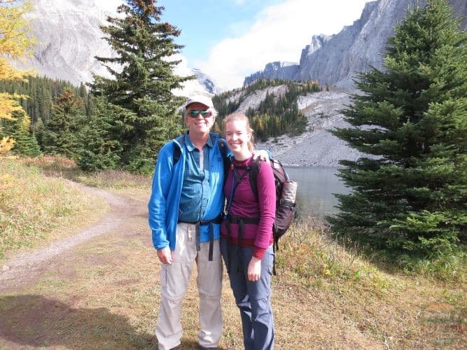 Man to the left is wearing white convertible pants with a blue fleece, hat and sunglasses. Lady to his right in a purple shirt and grey pants with mountains and a lake in the background.