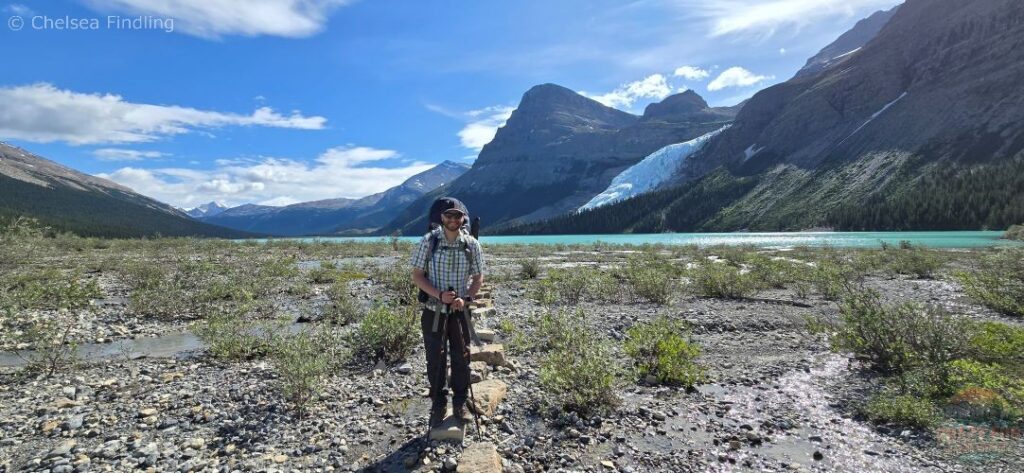 Male hiker standing on a rock along the lower trail with turquoise Berg Lake stretching out behind him.