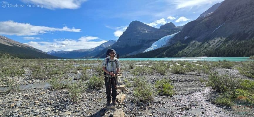 Man backpacking standing in front of the turquoise Berg Lake trail with mountains in the backdrop.
