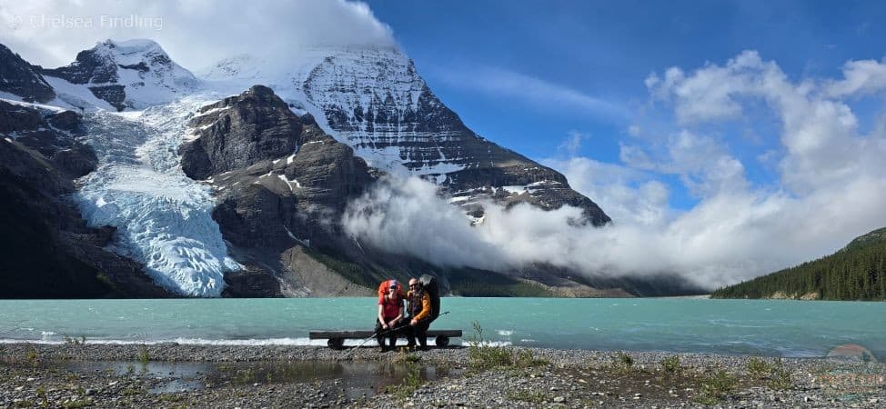 Two people seated on a wooden bench with the towering peak of Mt. Robson in the distance.