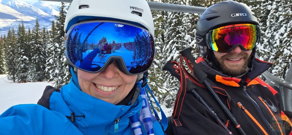 Two skiers on a chairlift with snowy mountains ahead, riding up to the peak at Marmot Basin.
