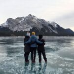 Back view of three women standing on the frozen bubble surface of Abraham Lake, AB, facing the Rocky Mountains in the background.