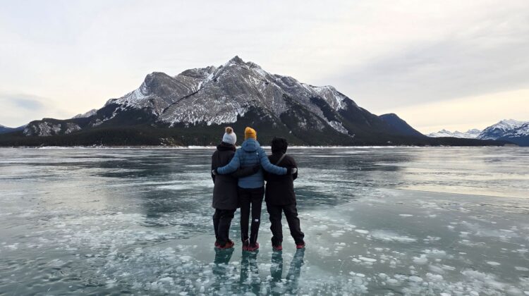 Back view of three women standing on the frozen bubble surface of Abraham Lake, AB, facing the Rocky Mountains in the background.