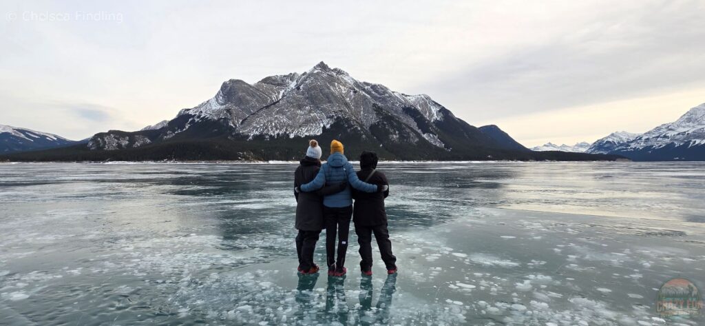 The backs of three women standing on the frozen surface of Abraham Lake, looking toward the mountains, with methane bubbles visible beneath the clear ice.