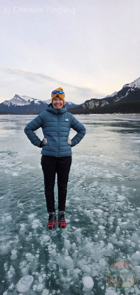 Woman standing on the frozen surface of Abraham Lake, surrounded by clusters of trapped methane ice bubbles visible beneath the clear blue ice.