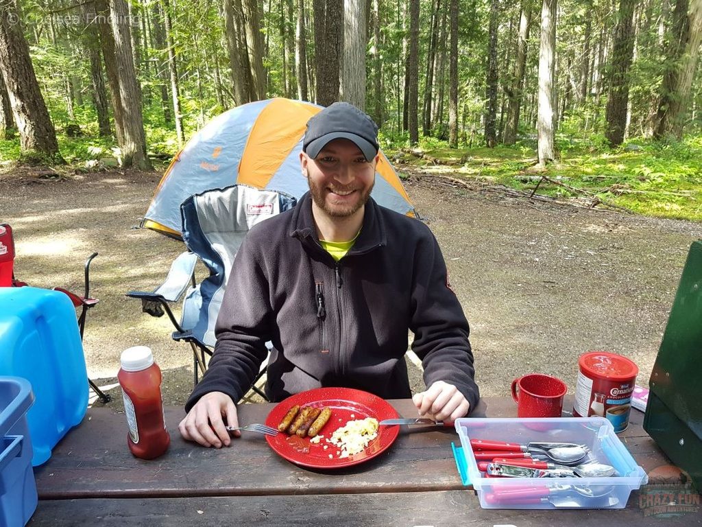 Man eating breakfast at a picnic table with eggs and sausage on a plate, enjoying a meal outdoors.