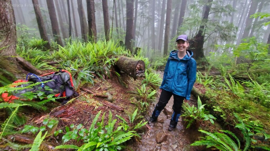 A backpacker taking a break on the muddy West Coast Trail, standing beside her pack in a damp coastal forest.
