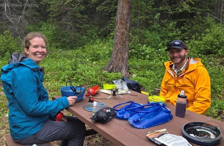 Two hikers enjoying warm backpacking meals for supper at a picnic table on the Berg Lake Trail.