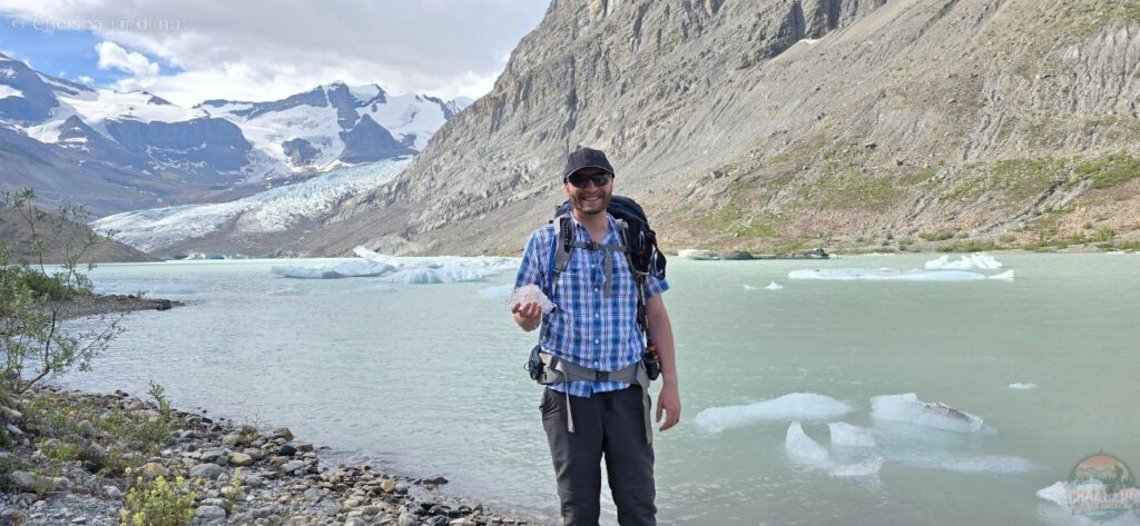 A man wearing a baseball cap holds a piece of ice beside an iceberg-filled lake, highlighting the need for warm layers like a toque and mitts in cold weather.