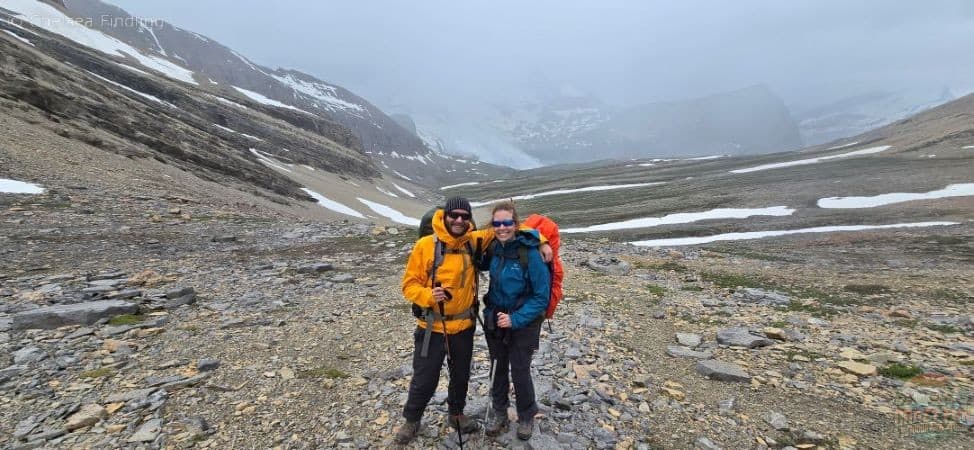 Two backpackers climbing Snowbird Pass as storm clouds roll in, carrying essentials from a backpacking gear list for a mountain adventure.