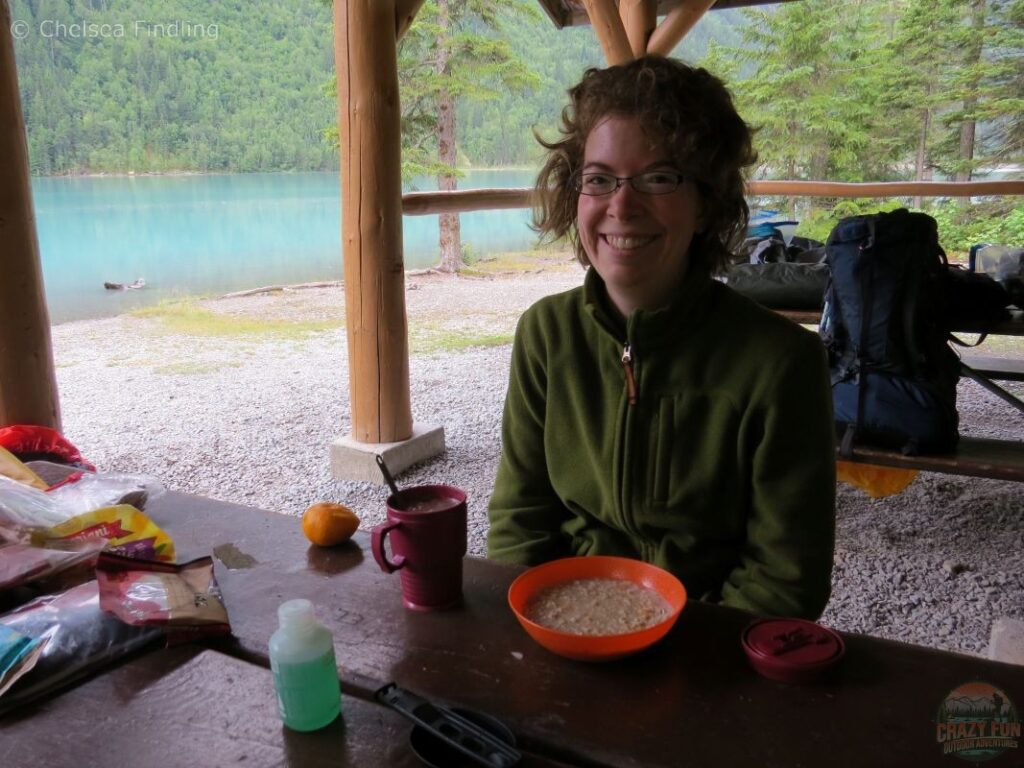A woman with messy hair eating breakfast at a picnic table while preparing her backpacking gear list before a big day on the trail.